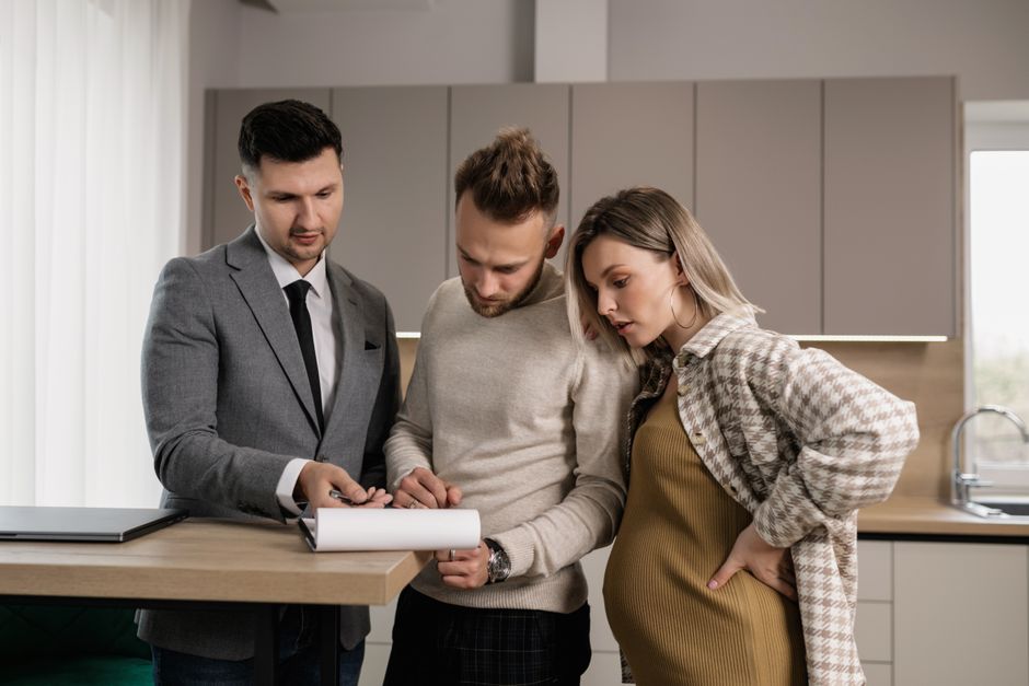 A young couple reviews and signs real estate documents with an agent in a modern kitchen setting.